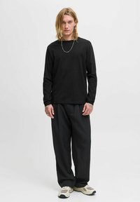 Langärmliges schwarzes Shirt kombiniert mit schwarzen Nadelstreifen-Hosen, die einen lässigen Schnitt aufweisen und mit einem grauen Sneaker-Design ausgestattet sind. Ein silberner Kettenakzent ist sichtbar.