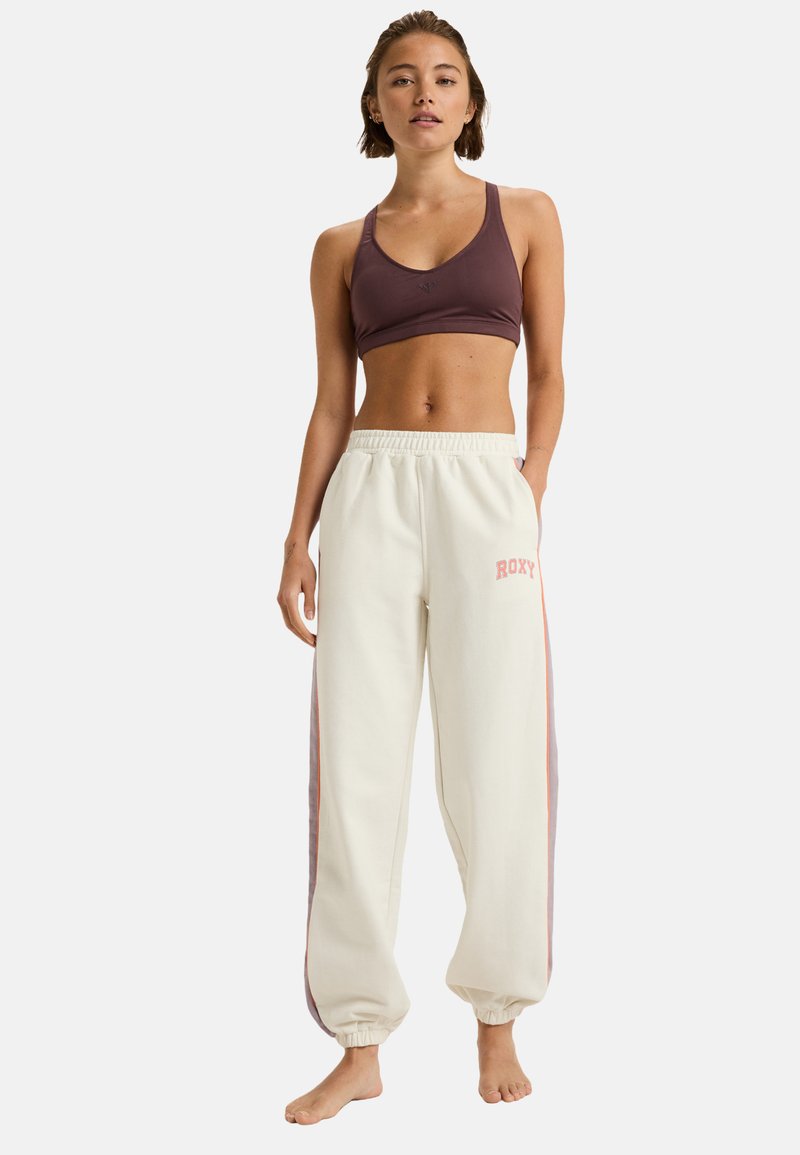 Sportbeha in donker mauve en crèmekleurige joggingsbroeken met roze zijkanten en het "ROXY" logo. Zachte, comfortabele stof met een elastische tailleband.