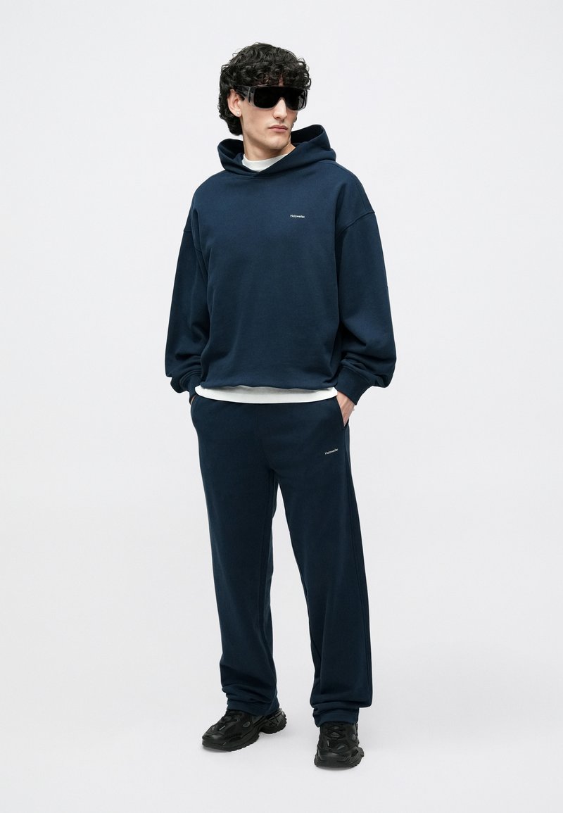 Navy blue cotton hoodie with kangaroo pocket and matching sweatpants. Black sunglasses and textured black sneakers complete the outfit.