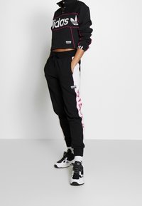 Svart croppad sweatshirt med en framträdande vit "adidas"-logotyp, ihop med svarta joggers med rosa och grå sidopaneler. Svartvita sneakers.