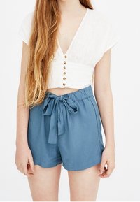 Top bianco a maniche corte con scollatura a V profonda e bottoni dorati, abbinato a pantaloni corti azzurri chiari a vita alta con un nodo frontale.