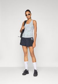 Top de alças azul claro com padrão, calções de desporto pretos com bolsos laterais, meias brancas e ténis pretos. Acessórios incluem uma mochila preta e óculos de sol.