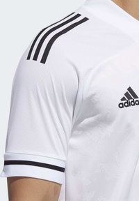 Hvid atletisk skjorte med en struktureret stof, sort tre-stribe design på skulderen og et sort logo på brystet.