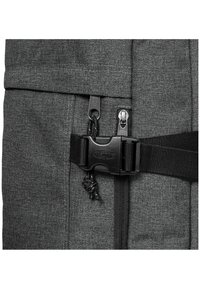 Šedý látkový batoh s černým uzávěrem na sponu, dvojitými zipy a strukturovaným povrchem se viditelnými švy.