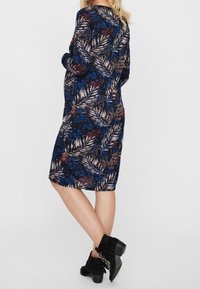 Langärmliges, marineblaues Kleid mit einem tropischen Blattmuster in Blau, Beige und Burgunderrot. Das Model kombiniert es mit schwarzen Knöchelstiefeln.
