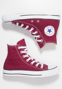 Wysokie trampki z burgundowego płótna, z białymi sznurówkami, czarnym wykończeniem i gumową podeszwą; mają naszywkę z logo Converse na kostce.