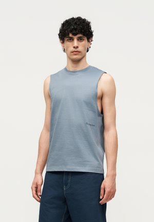 Νεαρός άνδρας με σγουρά σκούρα μαλλιά που φοράει αμάνικο ανοιχτό μπλε μπλουζάκι και σκούρο μπλε παντελόνι σε απλό ανοιχτό φόντο.