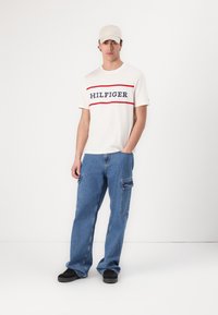 Cream T-shirt with navy and red "HILFIGER" logo, paired with loose blue denim cargo pants, and a light beige cap. Black shoes complete the look.
