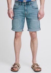 Shorts en denim bleu clair avec des revers roulés, comportant cinq poches et une légère décoloration. Portés avec des sandales brunes à lanières.