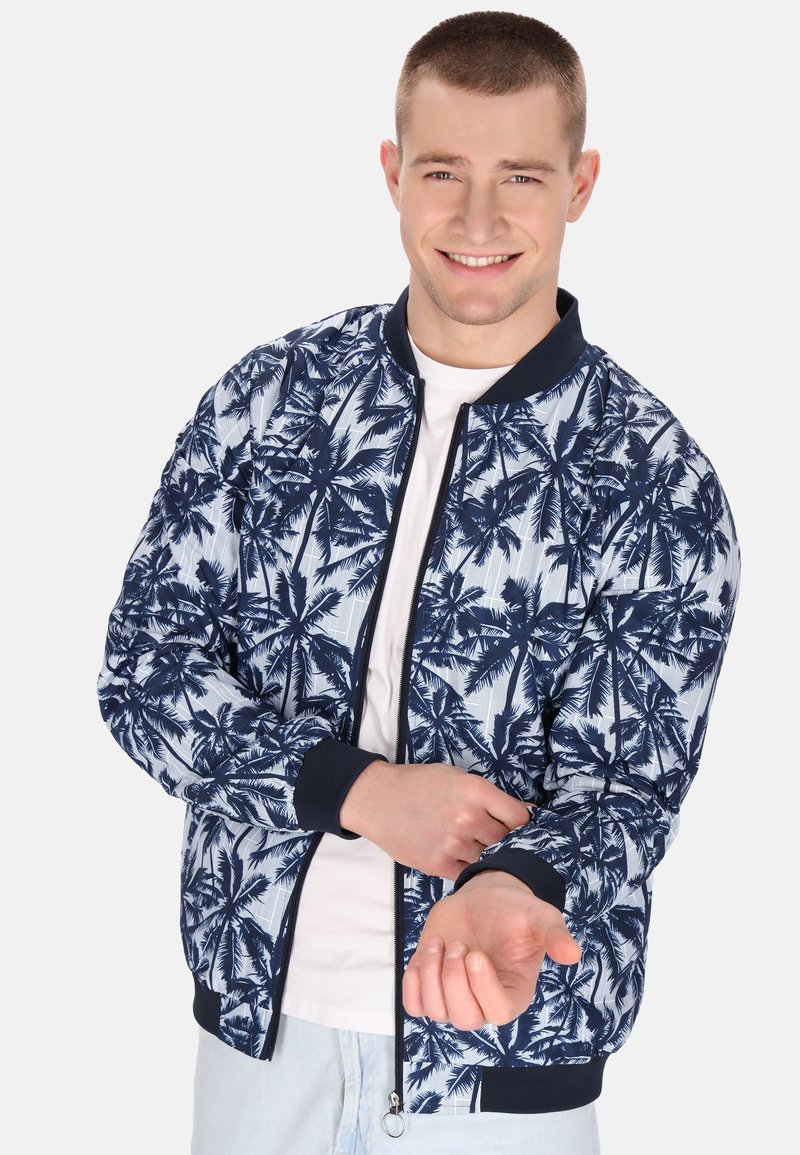 Marineblå og lyseblå bomberjakke med palmebladmønster, ribbede manchetter, lynlås lukning, båret over en hvid t-shirt. Lyse denimbukser.