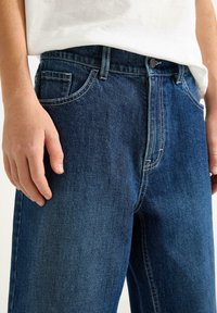 Mörkblå denimjeans med en klassisk femficksdesign, synlig söm och dragkedja med knappstängning. Slät textur.
