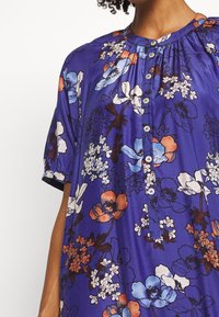 Vrouw die een paarse bloemenblouse draagt met blauwe, witte en oranje bloemen en een knoopsluiting aan de voorkant met korte pofmouwen.