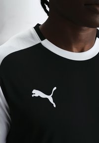 Mann trägt schwarz-weißes Sportshirt mit Puma-Logo auf der Brust, zeigt Hals und teilweise Gesicht vor einfachem Hintergrund.