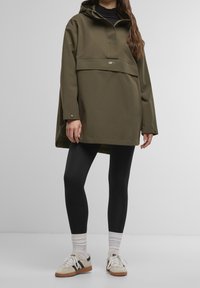 Personne portant une veste à capuche vert olive oversize, des leggings noirs, des chaussettes blanches et des baskets blanches avec des accents noirs.