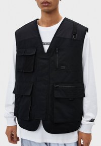 Chaleco negro con múltiples bolsillos, hecho de tela resistente, que cuenta con varios bolsillos, una cremallera frontal y un diseño sencillo sin cierres.