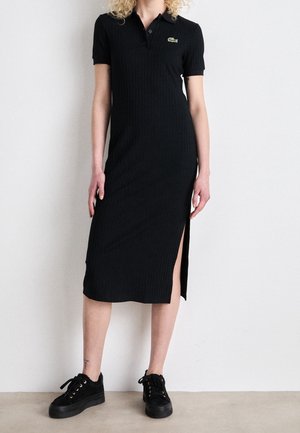 Femme portant une robe polo midi noire côtelée avec une fente latérale et des baskets plateformes noires, debout contre un mur clair uni.