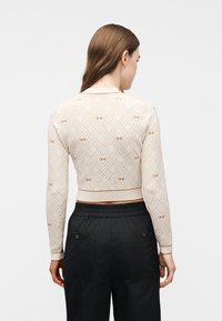 Γυναίκα που φαίνεται από πίσω, φοράει ένα μπεζ cropped πουλόβερ με μακριά μανίκια και σχέδιο, και φαρδιά μαύρη παντελόνια μπροστά σε απλό φόντο.