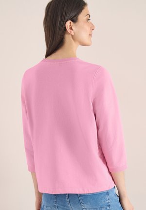 Femme aux longs cheveux foncés portant un haut rose clair à manches longues et un jean bleu, de profil sur un fond beige uni.