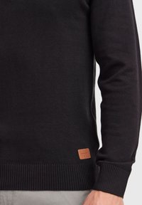 Pull en tricot noir avec des poignets et un ourlet côtelés. Présente un tissu texturé et une petite étiquette logo en cuir brun en bas à gauche.