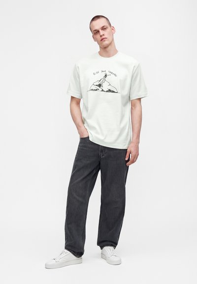 Νέος άνδρας που φοράει λευκό T-shirt με το κείμενο και το γραφικό "Ride Your Iceberg", μαύρο χαλαρό τζιν, λευκά αθλητικά, στέκεται με το ένα χέρι στην τσέπη.
