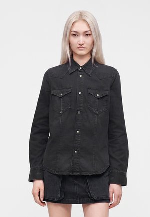 Jeune personne aux longs cheveux blonds portant une chemise en jean noire boutonnée et une jupe en jean noire assortie sur un fond uni.