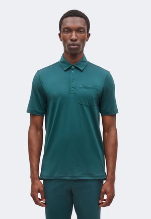Homme portant un polo turquoise à manches courtes avec une poche poitrine boutonnée, se tenant devant un fond clair uni.