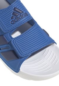 Modra atletska sandal s teksturiranimi sintetičnimi trakovi, ki vsebuje logo, nastavljivo zapiranje s hook-and-loop ter belo oblazinjeno podplato.