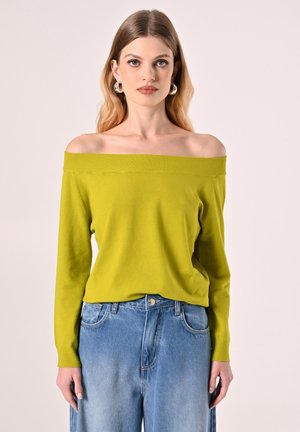 Limoengroene off-shoulder trui van gebreide stof, lange mouwen, geribbelde halslijn. Gecombineerd met lichtblauwe highwaisted spijkerbroek.