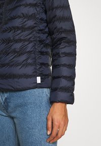 Person wearing a black puffer jacket and blue jeans, shown from mid-torso to hand against a plain light background.