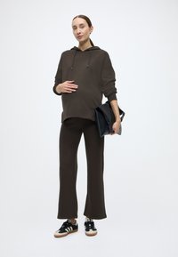 Καφέ φούτερ με κουκούλα και κορδόνι, με άνετη εφαρμογή, σετ με καφέ παντελόνι καμπάνα. Μαύρη τσάντα και αθλητικά παπούτσια με ανοιχτές λεπτομέρειες.