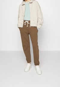Felpa con zip beige, dotata di corde di regolazione, abbinata a pantaloni felpati marroni a gamba affusolata con vita elastica e sneaker bianche.