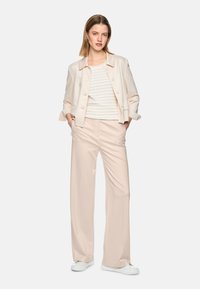 Woman standing wearing a light beige jacket, striped beige and white top, wide-leg beige pants, and white sneakers on a white background.