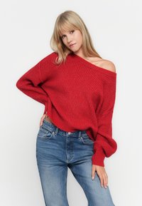 Un pull surdimensionné en tricot rouge avec un grand col et des manches longues, associé à un jean en denim bleu. La texture est épaisse et décontractée.