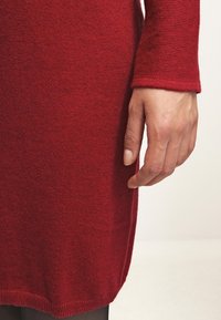 Robe tricotée rouge avec une finition texturée. Gros plan d'une main reposant sur l'ourlet, révélant de longues manches et des fentes sur les côtés en bas.