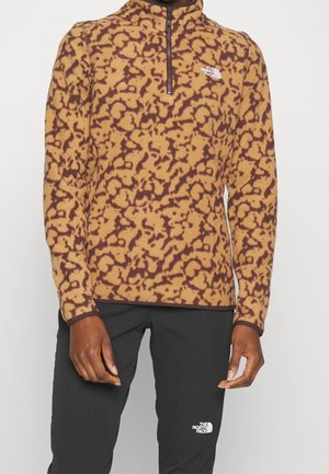 Mężczyzna ubrany w brązowo-beżową wzorzystą bluzę polarową z krótkim zamkiem i czarne spodnie, oba z logo The North Face.