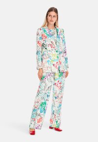 Blumenbedruckter Anzug in Weiß mit bunten Blatt- und Blütenmustern. Besteht aus einem taillierten Blazer und weit geschnittenen Hosen; kombiniert mit roten Schuhen.