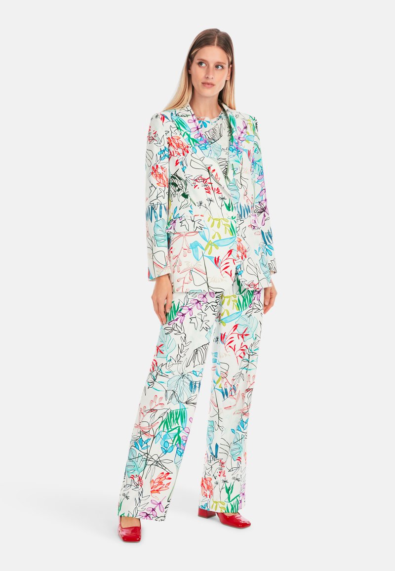 Blumenbedruckter Anzug in Weiß mit bunten Blatt- und Blütenmustern. Besteht aus einem taillierten Blazer und weit geschnittenen Hosen; kombiniert mit roten Schuhen.