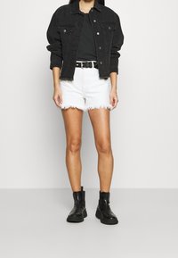 Chaqueta de mezclilla negra con dobladillo deshilachado, combinada con pantalones cortos blancos desgastados. Cinturón negro, botas negras hasta el tobillo y camiseta negra ajustada.