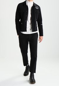 Zwarte spijkerjack met knopen aan de voorkant en een logo-opdruk op de borst, gedragen over een wit T-shirt, gecombineerd met zwarte tapering broek en zwarte enkellaarsjes.