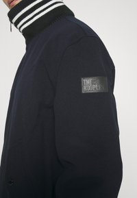 Personne portant une veste foncée avec un col montant rayé et un patch en cuir noir sur la manche libellé "THE KOOPLES."