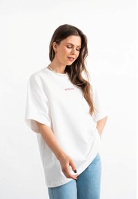 Biała, oversize koszulka z krótkim rękawem, zdobiona różowym haftowanym napisem "MIRAGE" na przodzie. Wykonana z miękkiego bawełnianego materiału, w luźnym kroju.