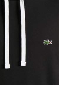 Felpa nera con lacci bianchi, caratterizzata da un piccolo logo ricamato in verde e bianco a forma di coccodrillo sul davanti. Materiale morbido.