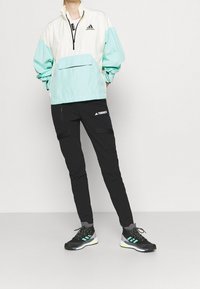 Chaqueta halter ligera con bloques de color que presenta un cierre de cremallera y un bolsillo frontal. Combinada con pantalones cargo negros y zapatillas deportivas con acentos neón.