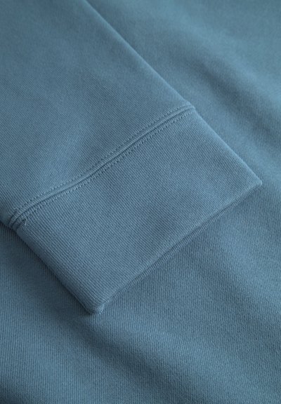 Țesătură de bumbac albastră pliată pentru a arăta un mânecă cu o manșetă cu model, având o textură netedă și detalii de cusătură curate.