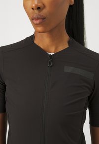 Maillot de cyclisme noir avec une fermeture éclair quart, manches courtes et tissu texturé. Présente un logo mat accent sur la poitrine.