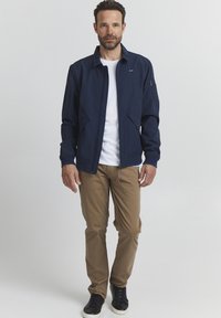 Navy blue jacket with a stand collar, two side pockets, and a zippered sleeve pocket. Worn over a white t-shirt and paired with tan pants and black sneakers.