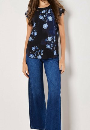 Kobieta w czarnej bluzce z niebieskimi kwiatowymi wzorami i szerokich, wysokich spodni dżinsowych z szerokimi nogawkami, na jednolitym tle.