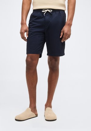 Άνδρας που φορά μπλε ναυτικού τύπου σορτς με κορδόνι και μπεζ παπούτσια χωρίς κορδόνια, στέκεται μπροστά σε απλό ανοιχτόχρωμο φόντο.