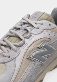 Gros plan d'une basket beige et grise avec tissu mesh, détails en suède, lacets gris et un logo réfléchissant "N" sur le côté.