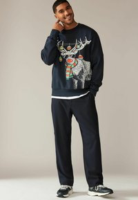 Felpa blu navy con un grafico di una renna con occhiali da sole, ornamenti colorati e una sciarpa a righe, abbinata a pantaloni scuri e sneaker.